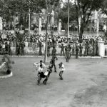 A row of soldiers keep a large crowd of people from coming through the fence into a plaza. In the center of the photograph, a soldier and a civilian carry an injured man. Another man takes a picture