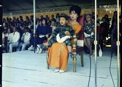 Madame Nhu sitting on a stage with her son, wearing a soldier's outfit, in her lap. A crowd of people sit behind her.