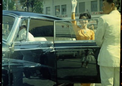 Madame Nhu waving as she exits her black car. Keever is reflected in the door of the car.