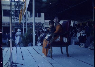 Madame Nhu sitting on a stage with her son, wearing a soldier's outfit, in her lap. A crowd of people sit behind her.