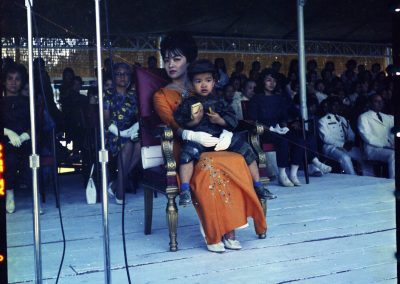 Madame Nhu sitting on a stage with her son, wearing a soldier's outfit, in her lap. Her son holds a little yellow box in one hand. A crowd of people sit behind her.