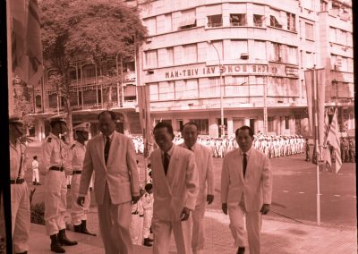 A group of men are walking into a building. Many men in uniform are seen standing on the street in the background.
