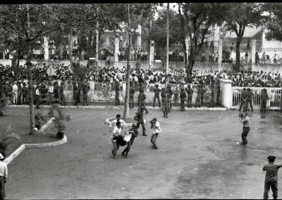 A row of soldiers keep a large crowd of people from coming through the fence into a plaza. In the center of the photograph, a soldier and a civilian carry an injured man. Another man takes a picture.