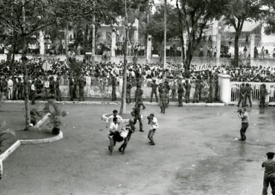 A row of soldiers keep a large crowd of people from coming through the fence into a plaza. In the center of the photograph, a soldier and a civilian carry an injured man. Another man takes a picture