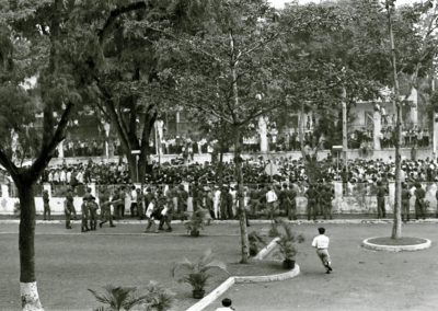 A row of soldiers keep a large crowd of people from coming through the fence into a plaza. To the left, a soldier and a civilian carry an injured man.