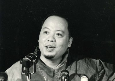 Khanh sitting at a press conference as his photo is taken. He appears to be responding to some type of question. Four microphones are seen in front of him.